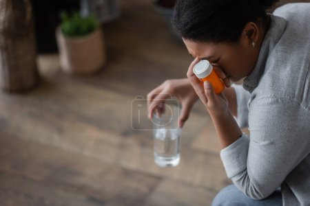Dissatisfied multiracial woman holding pills and blurred glass of water at home 