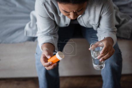 mujer multirracial estresada sosteniendo píldoras y agua mientras se sienta en la cama borrosa 