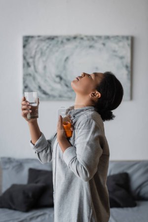 Téléchargez les photos : Vue latérale de la femme multiraciale prenant la pilule antidépresseur et tenant l'eau dans la chambre - en image libre de droit