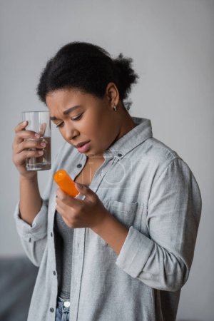 Upset multiracial woman holding pills and glass of water at home 