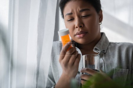 Traurige Frau mit verschwommenen Pillen und Wasser in der Nähe von Vorhang zu Hause 
