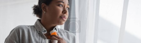 Depressed multiracial woman holding antidepressant pills near curtain at home, banner 