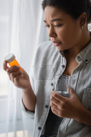 Mujer joven multirracial con depresión sosteniendo pastillas y vaso de agua en casa 