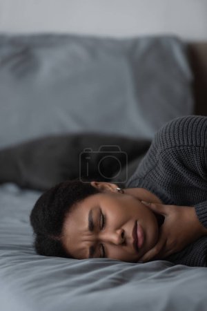 Sad multiracial woman with depression touching neck while lying on bed at home 