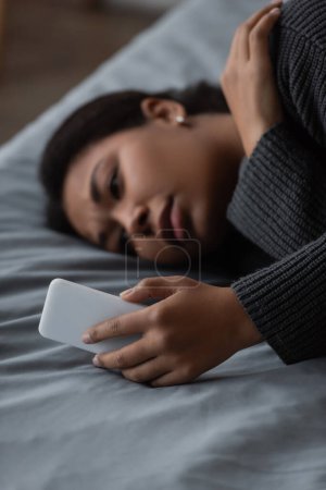 Verschwommene multiethnische Frau mit Depressionen nutzt Smartphone, während sie zu Hause im Bett liegt 