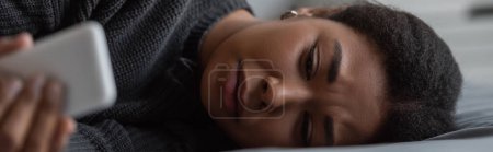 Mujer joven multirracial con depresión usando teléfono inteligente en la cama, pancarta 