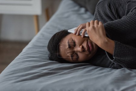 Depressed multiracial woman calling helpline while lying on bed at home 
