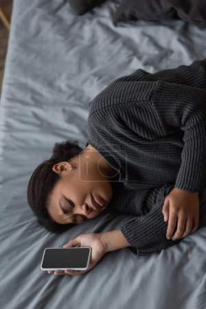 Draufsicht der unzufriedenen multirassischen Frau im Pullover, die ihr Handy auf dem Bett hält 