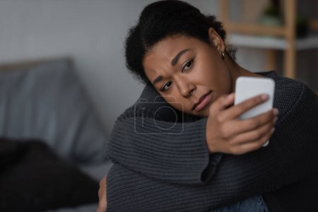 heartbroken multiracial woman in sweater using blurred smartphone in blurred bedroom 