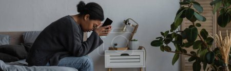 Téléchargez les photos : Vue latérale d'une femme multiraciale déprimée tenant un téléphone portable alors qu'elle était assise sur son lit à la maison, bannière - en image libre de droit