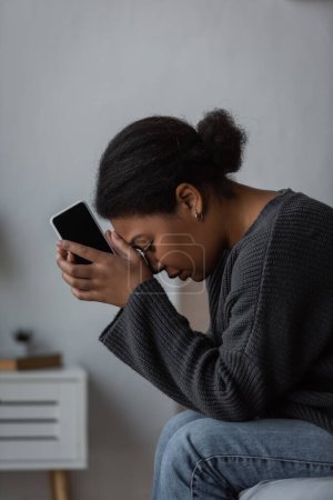 Foto de Vista lateral de la joven mujer multirracial con problemas mentales sosteniendo el teléfono celular mientras está sentada en la cama en casa - Imagen libre de derechos
