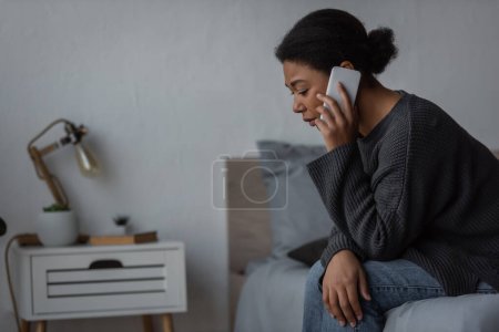 heartbroken multiracial woman in sweater talking on smartphone on bed at home 