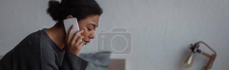 Mujer multirracial molesta en suéter hablando en el teléfono celular en el dormitorio en casa, pancarta 
