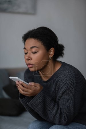 Foto de Mujer joven multirracial usando teléfono inteligente mientras está sentado en la cama borrosa en casa - Imagen libre de derechos