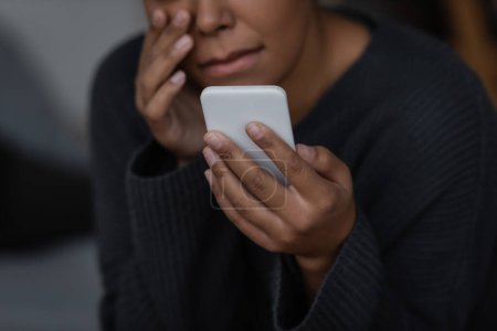 Verschwommene Ansicht einer multiethnischen Frau mit Depressionen, die ihr Smartphone zu Hause hält 