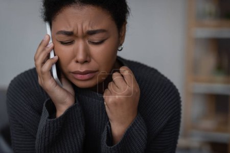 Crying multiracial woman talking on smartphone with helpline at home 