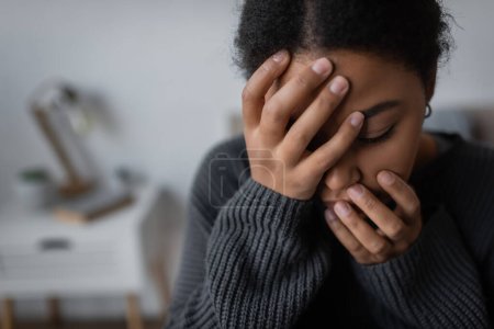 Junge multiethnische Frau mit Depressionen berührt Gesicht in verschwommenem Haus 