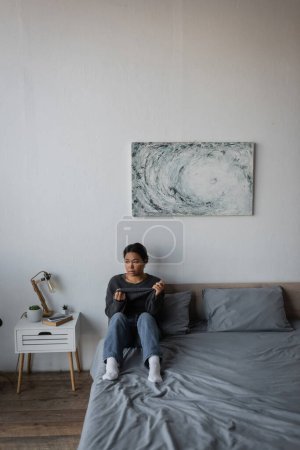 Frustrated multiracial woman touching sweater while sitting on bed at home 