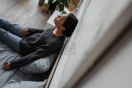 Blick aus der Vogelperspektive auf eine traurige multiethnische Frau in Strickpullover, die an der Wand lehnt, während sie zu Hause auf dem Bett sitzt 