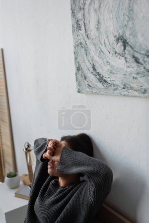 Photo for Depressed multiracial woman in knitted sweater covering eyes in bedroom - Royalty Free Image