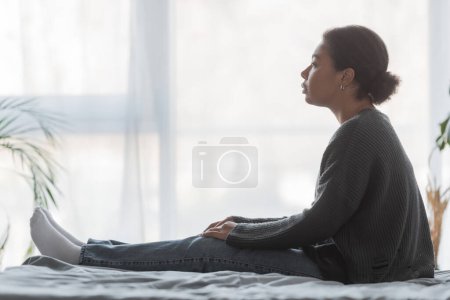 Téléchargez les photos : Vue latérale de la femme multiraciale avec apathie regardant loin tout en étant assis sur le lit à la maison - en image libre de droit