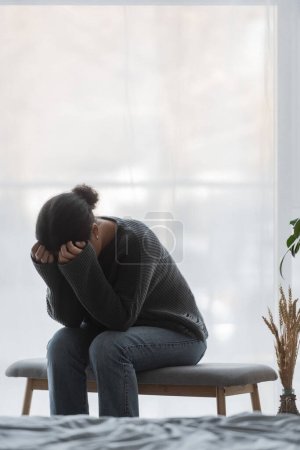 mujer multirracial con el corazón roto cubriendo la cara mientras está sentado en el banco de la cama en casa 
