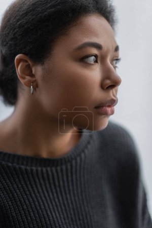 Frustrated multiracial woman in grey knitted sweater looking away at home 