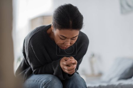 Depressed multiracial woman in knitted sweater looking at hands in bedroom 
