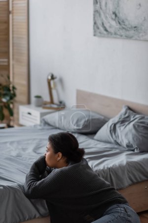 Foto de Mujer joven multirracial con problemas psicológicos mirando hacia otro lado cerca de la cama en casa - Imagen libre de derechos