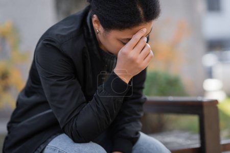 Foto de Deprimida mujer multirracial con chaqueta sentada en un banco borroso al aire libre - Imagen libre de derechos