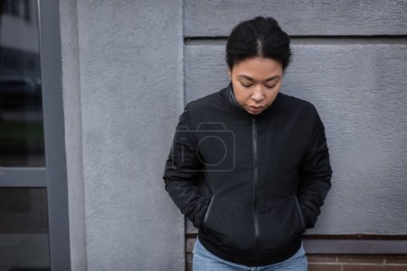 multiracial woman with mental problem looking down while standing near building on urban street 