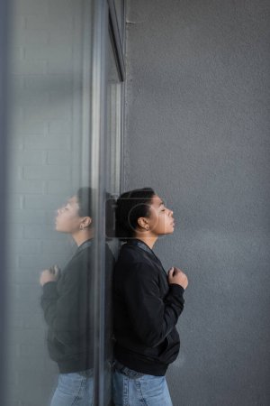 Téléchargez les photos : Vue latérale de la femme multiraciale avec dépression debout près de la façade du bâtiment à l'extérieur - en image libre de droit