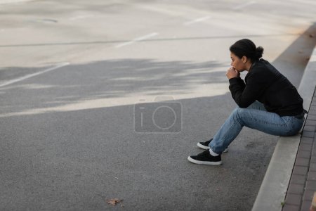 Téléchargez les photos : Vue de côté de la jeune femme multiraciale avec un problème mental assis à la frontière sur la rue urbaine - en image libre de droit