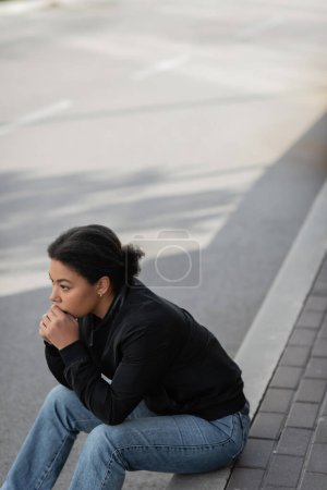 Junge multiethnische Frau mit Depressionen schaut weg, während sie an der Grenze im Freien sitzt 