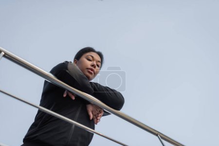 Bottom view of multiracial woman with depression looking at camera near railing on urban street 