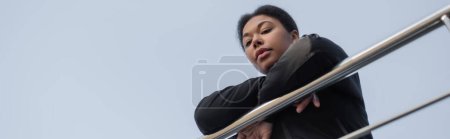 Bottom view of depressed multiracial woman looking at camera near railing outdoors, banner 