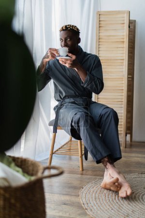 African american man with vitiligo in robe drinking coffee on bedroom bench at home 