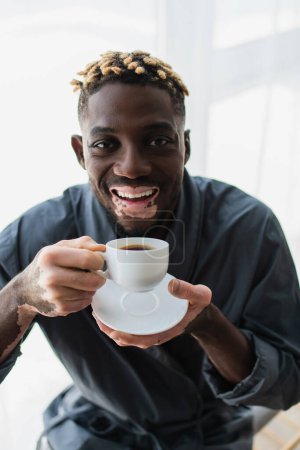 Porträt eines fröhlichen afrikanisch-amerikanischen Mannes mit Vitiligo, der zu Hause eine Tasse Kaffee hält 