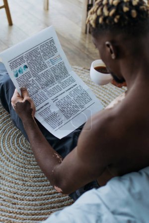 Foto de Hombre afroamericano sin camisa con vitiligo leyendo periódico y tomando café en casa - Imagen libre de derechos