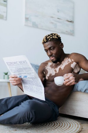Muskulöser afrikanisch-amerikanischer Mann mit Vitiligo, der im Schlafzimmer Kaffee hält und Zeitung liest 