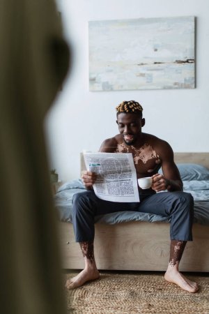 Photo for Smiling african american man with vitiligo reading news and holding coffee on bed at home - Royalty Free Image