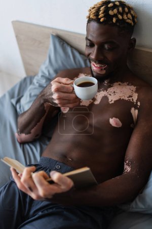 Hemdloser afrikanisch-amerikanischer Mann mit Vitiligo lächelnd, während er Kaffee hält und Buch liest 