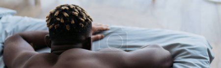 Back view of shirtless african american man with vitiligo relaxing on bed at home, banner
