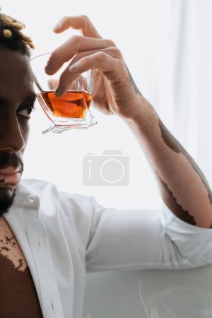 Cropped view of african american man with vitiligo holding whiskey while sitting in bathtub 