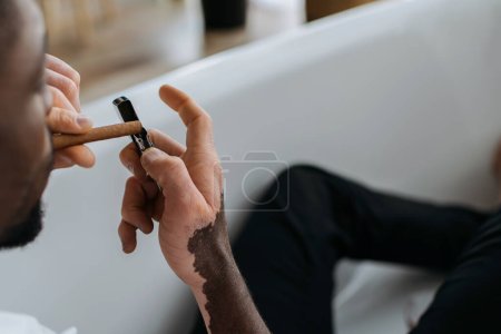 Blurred african american man with vitiligo holding lighter and cigar in bathtub 