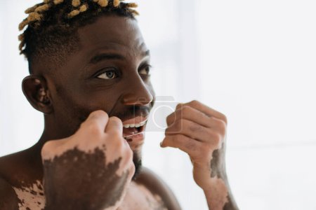 Téléchargez les photos : Sans chemise homme afro-américain avec vitiligo à l'aide de fil dentaire à la maison - en image libre de droit