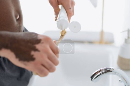 Foto de Vista recortada del hombre afroamericano con vitiligo apretando pasta de dientes en el cepillo de dientes en el baño - Imagen libre de derechos