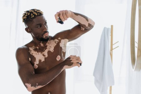 Shirtless african american man with vitiligo using deodorant near mirror in bathroom
