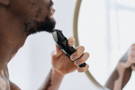 Cropped view of blurred african american man with vitiligo shaving with electric razor in bathroom 