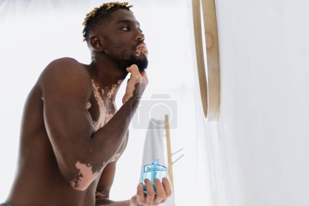 Low angle view of shirtless african american man with vitiligo applying lotion on face in bathroom 